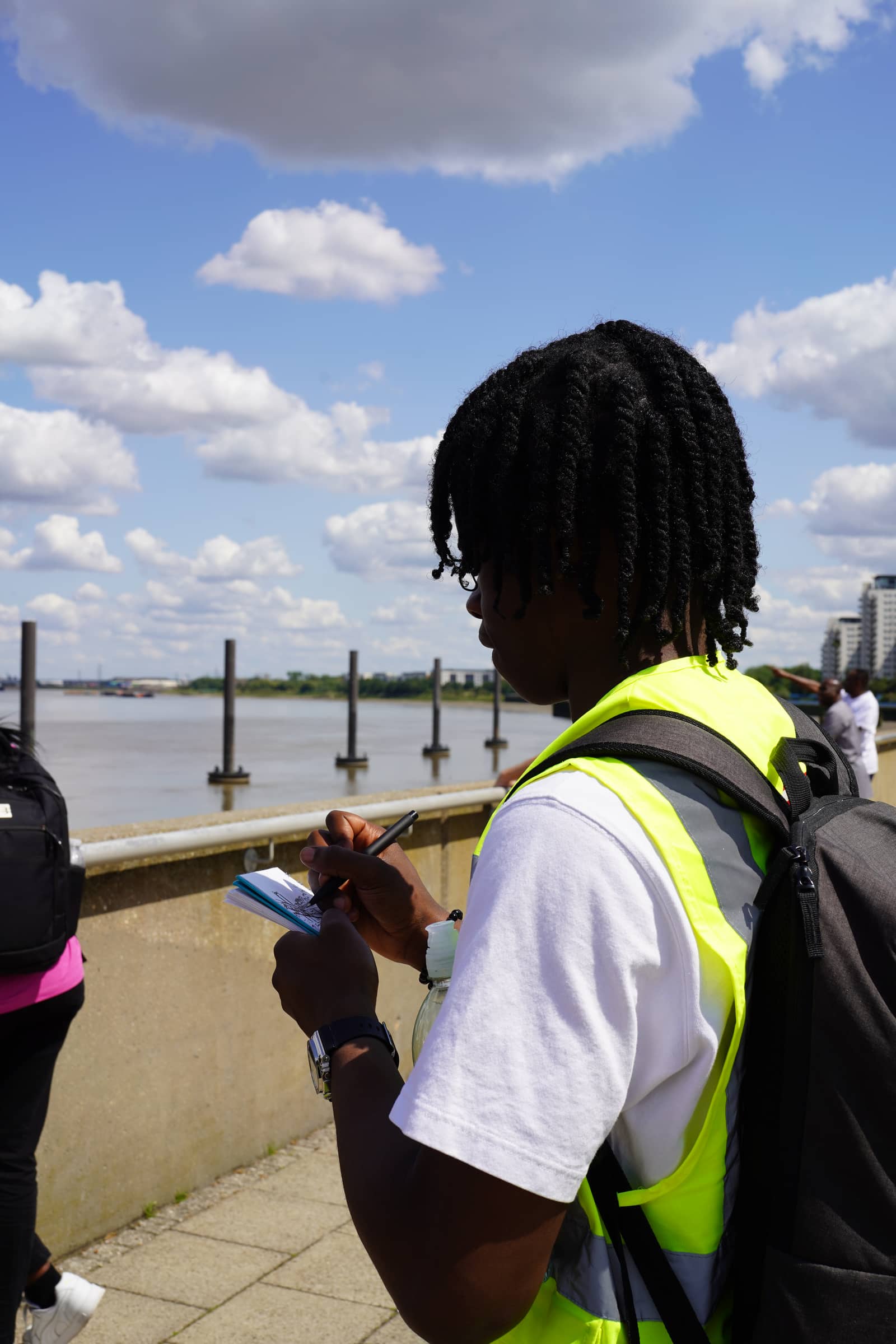 A young person in a hi-vis vest drawing in a notebook on the river path